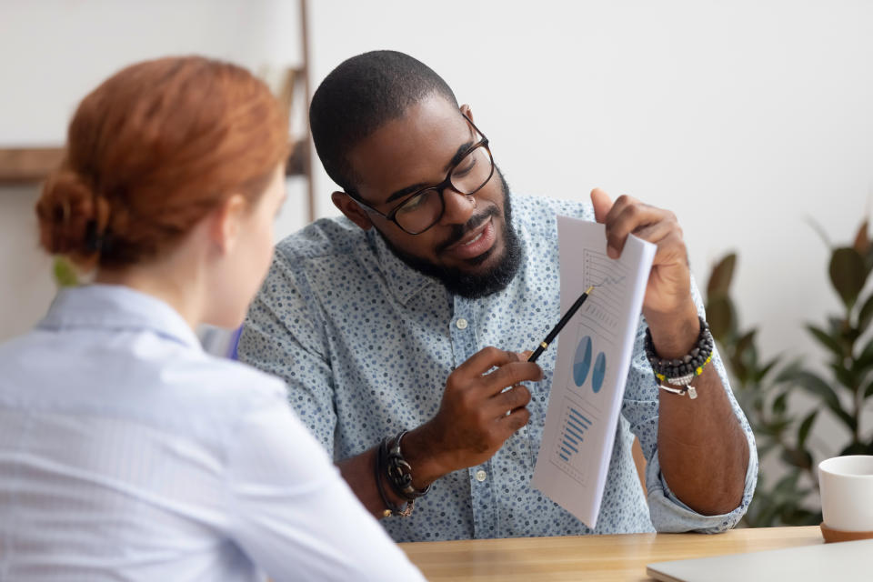 Male black coworker showing document with project results talking to caucasian female colleague. Diverse businesspeople analyzing consulting discussing project results in charts and graphs in office