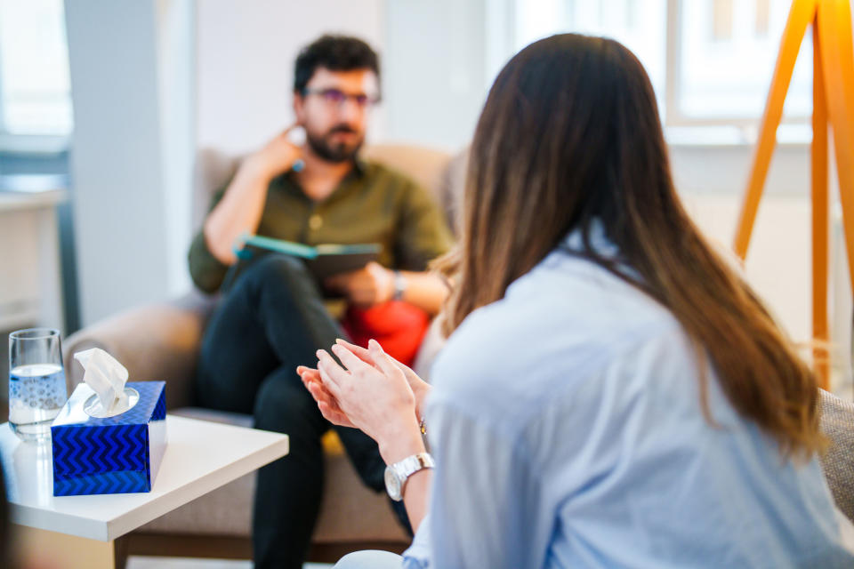 Two people in a professional meeting; one is talking while the other takes notes. A table with tissues and a glass of water is between them