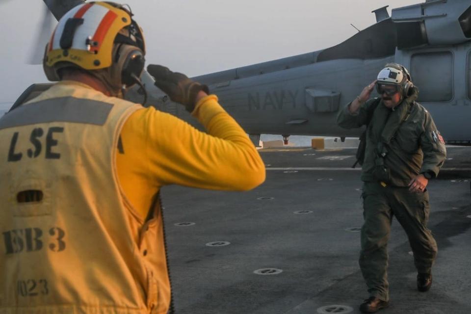 a military captain salutes a flight deck officer with a helicopter behind him