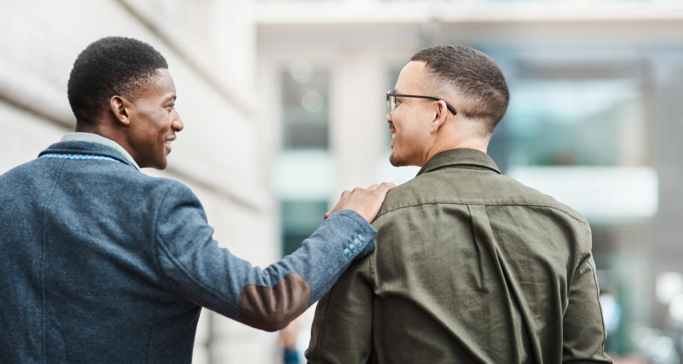 Two men, one in a blazer and the other in a casual shirt, smiling and talking while walking, with one man patting the other on the shoulder