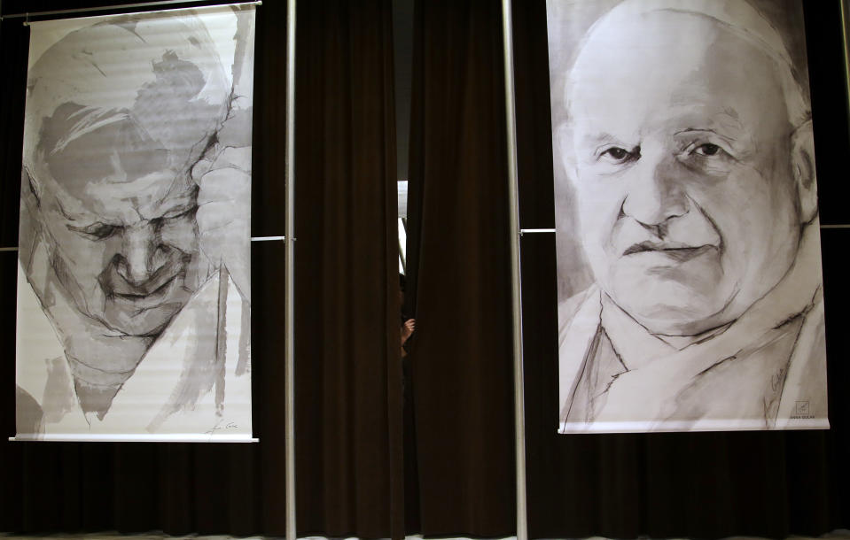 A press hall attendant holds the curtains adorned with portraits of late Pope John Paul II, left, and John XXIII prior to the start of a press conference at the Vatican, Friday, April 25, 2014. Hundred thousands of pilgrims and faithful are expected to reach Rome to attend the scheduled April 27 ceremony at the Vatican in which Pope Francis will elevate in a solemn ceremony John XXIII and John Paul II to sainthood. (AP Photo/Gregorio Borgia)
