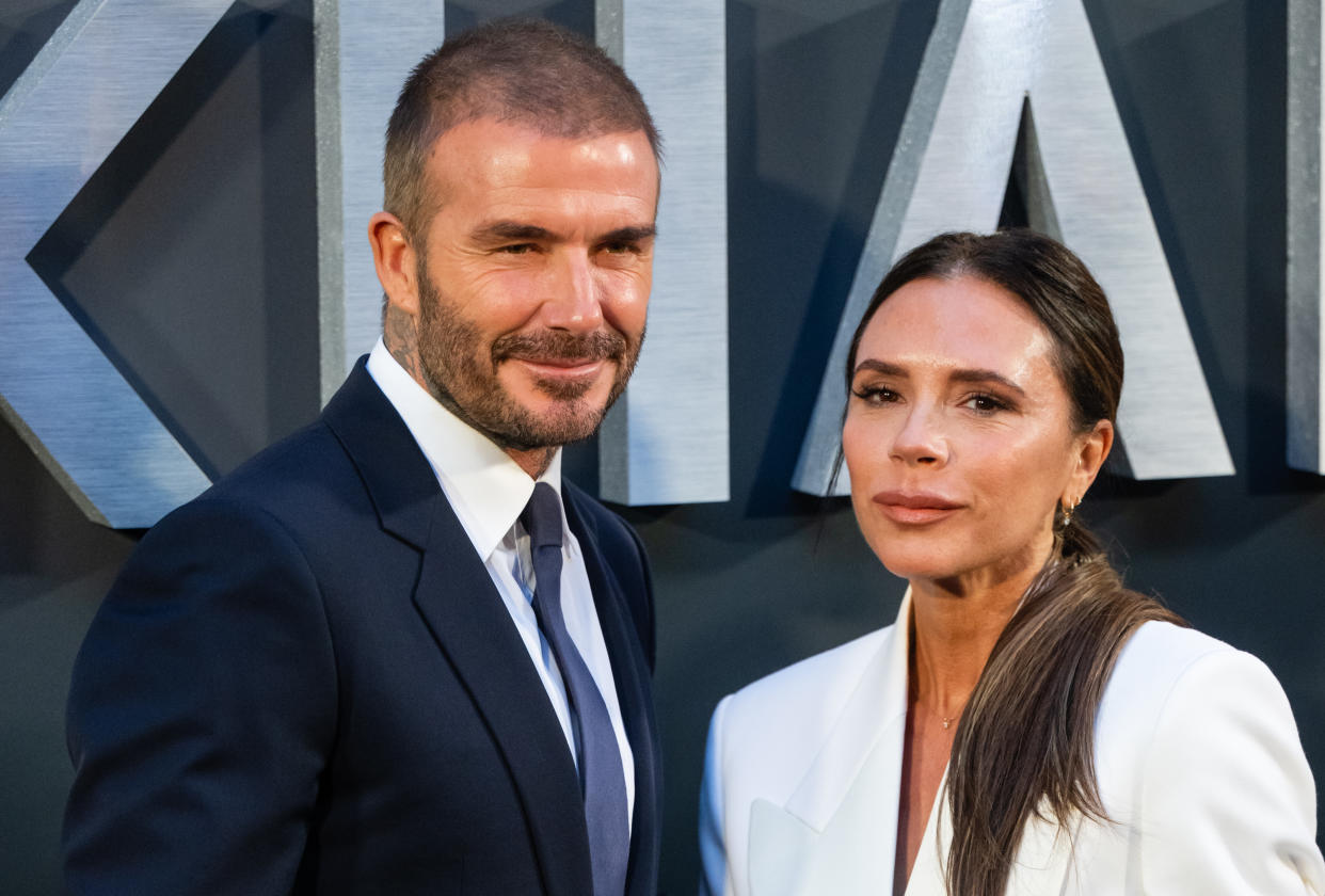 David Beckham, wearing a suit and tie, smiles as he stands next to Victoria Beckham, wearing a white blazer, on the red carpet