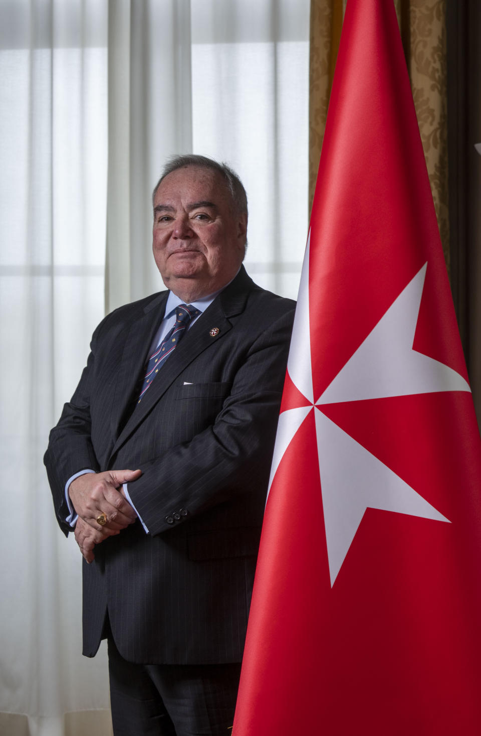 John Dunlap poses with the Sovereign Military Order of Malta's flag in the Chapter Room at their headquarters in Rome, Friday, March 31, 2023. Dunlap, a Canadian lawyer who found his vocation ministering to AIDS patients in Harlem has been elected Wednesday, May 3, 2023, the grand master of the Knights of Malta, the first non-European and first non-aristocratic head of the ancient Catholic order that provides humanitarian aid around the world. (AP Photo/Domenico Stinellis)