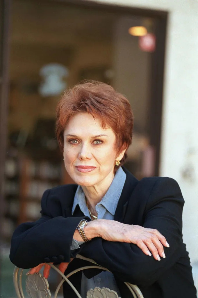 A woman with short hair leans on a chair, wearing a blazer over a collared shirt. Indoor setting with furniture in the background