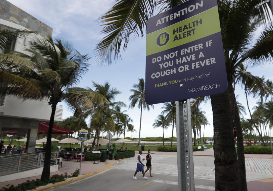 FILE - In this June 22, 2020, file photo a couple walks past a sign asking people not to visit Miami Beach, Florida's famed South Beach if they have a cough or fever. An Associated Press analysis of coronavirus case data shows the virus has moved, and is spreading quickly, into Republican areas, a new path with broad potential political ramifications. (AP Photo/Wilfredo Lee, File)