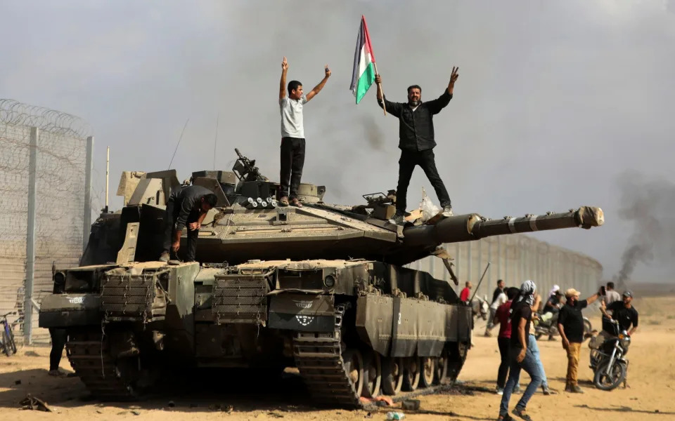 Palestinians wave their national flag and celebrate by a destroyed Israeli tank at the Gaza Strip fence east of Khan Younis