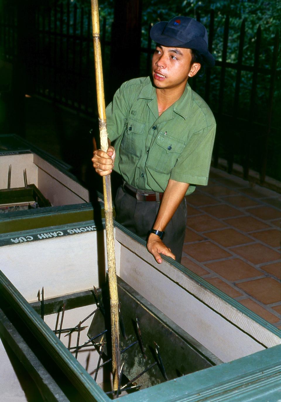 A photo of a man holding a stick through a trap door filled with long metal spikes.