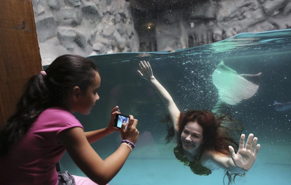 A woman dressed as a siren performs from inside a tank as school girls uses her mobile phone to take a photo of her at Sao Paulo Aquarium