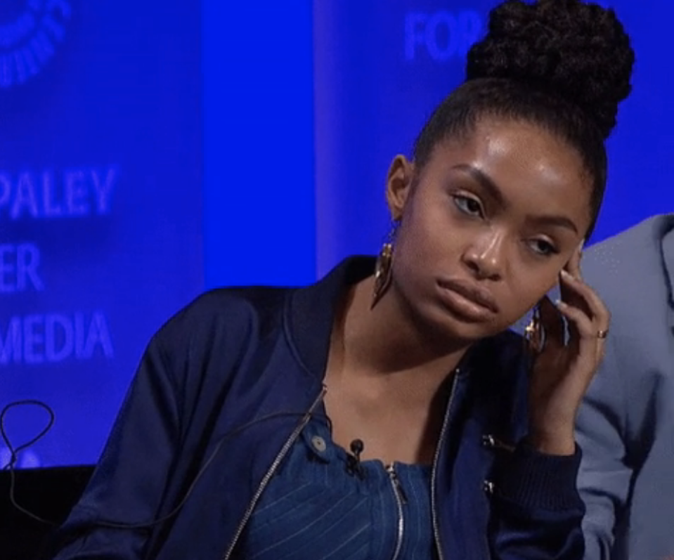 A woman with a top bun and hoop earrings looks thoughtful with her hand on her cheek