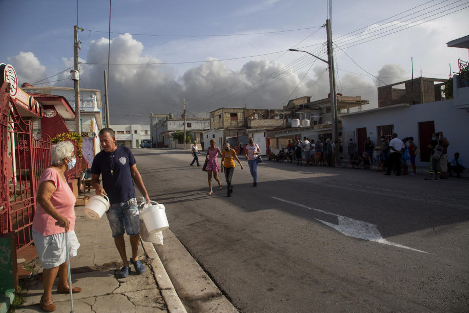 Residents walk in Matanzas, Cuba, Tuesday, Aug. 9, 2022 as smoke rises in the background from a deadly fire at a large oil storage facility. The fire was triggered by lighting that struck one of the facility’s eight tanks late Friday, Aug. 5th. (AP Photo/Ismael Francisco)