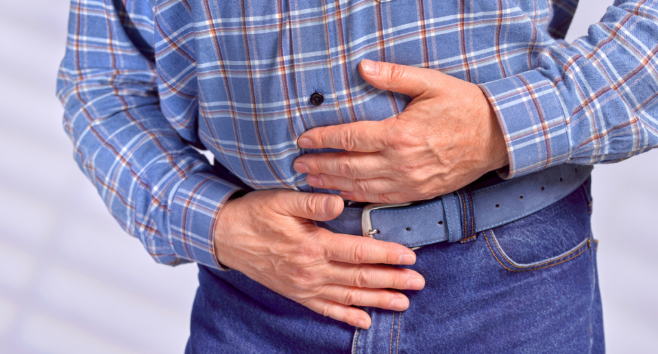 old man clutching stomach with hands wearing blue jeans and plaid shirt, colon cancer