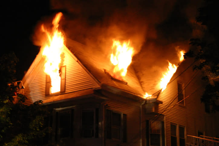 A house is engulfed in flames at night, with fire coming out from multiple windows and the roof. No people are visible