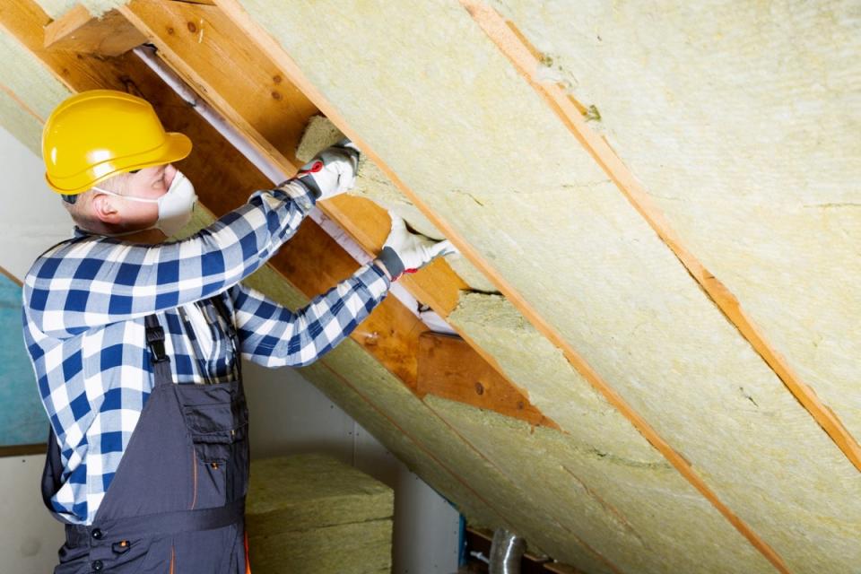 Man wearing a yellow hard hat and face mask installing insulation in attic.