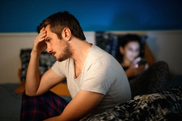 A man holds his head in frustration while his wife sits in bed and uses her phone