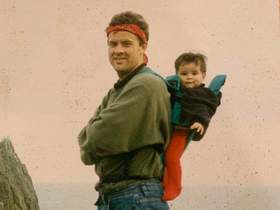 A man wearing a bandanna stands on a cliff in the 1990s with a baby on his back in a carrier.