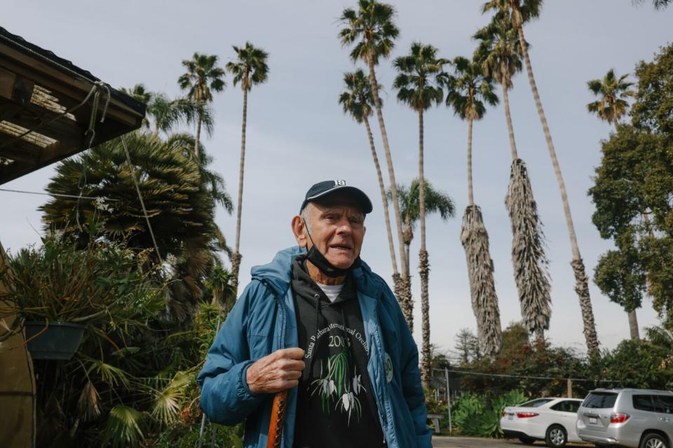 A man with a face mask under his chin stands outside a nursery, with palm trees in the background