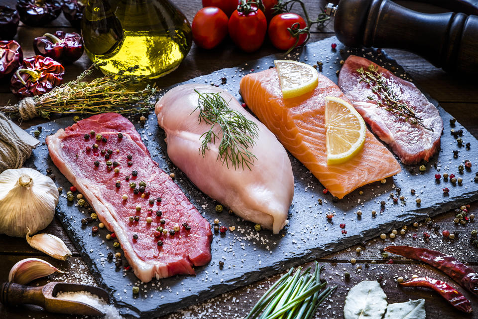 High angle view of four different types of animal protein like a raw beef steak, a raw chicken breast, a raw salmon fillet and a raw pork steak on a stone tray surrounded by condiments, spices and vegetables. Low key DSLR photo taken with Canon EOS 6D Mark II and Canon EF 24-105 mm f/4L