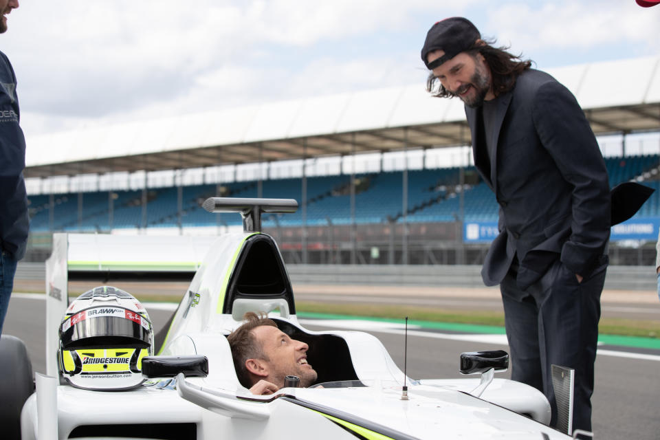 Jenson Button sits in a Formula One car while Keanu Reeves stands next to him, looking down.