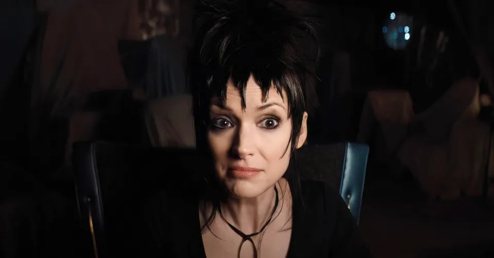 Winona Ryder as Lydia Deetz in "Beetlejuice" revival, sporting a gothic outfit with a black choker and spiky hairstyle, expressing a perplexed look