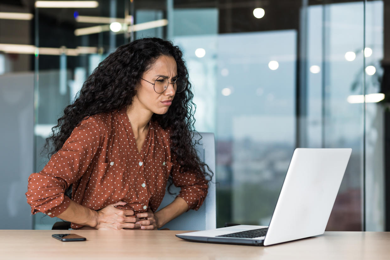 Latin American business woman is sick at work, employee with curly hair and glasses has severe stomach pain, businesswoman working inside office building using laptop