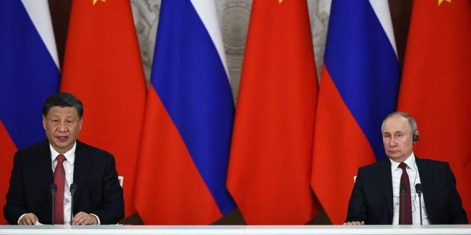 Xi Jinping and Vladimir Putin sitting at a conference table with Russian flags in the background.