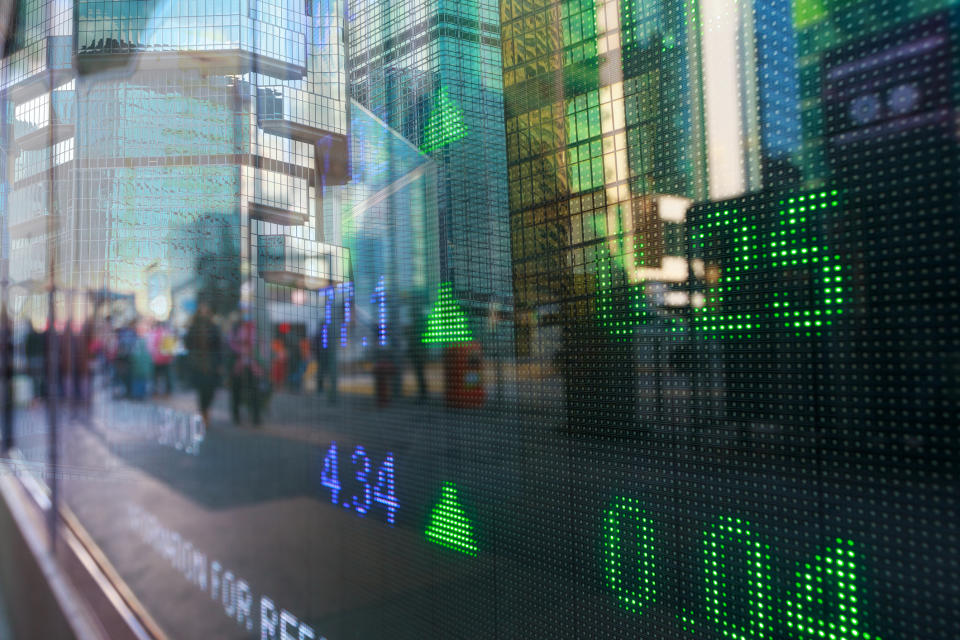 Stock market prices on a digital window display with city reflection on the glass.