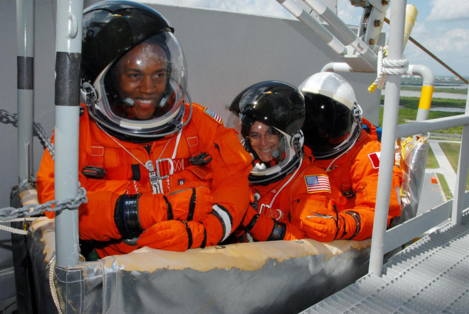 three people stand in a stretcher in bright orange spacesuits.