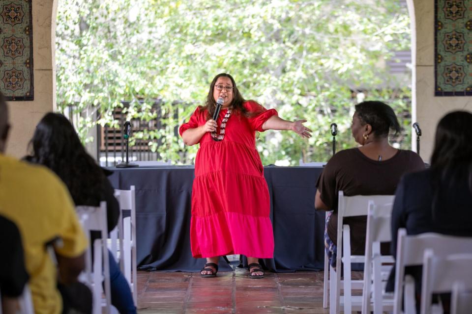 A woman in long, red dress stands at the center, speaks in a microphone before an audience seated on both sides.