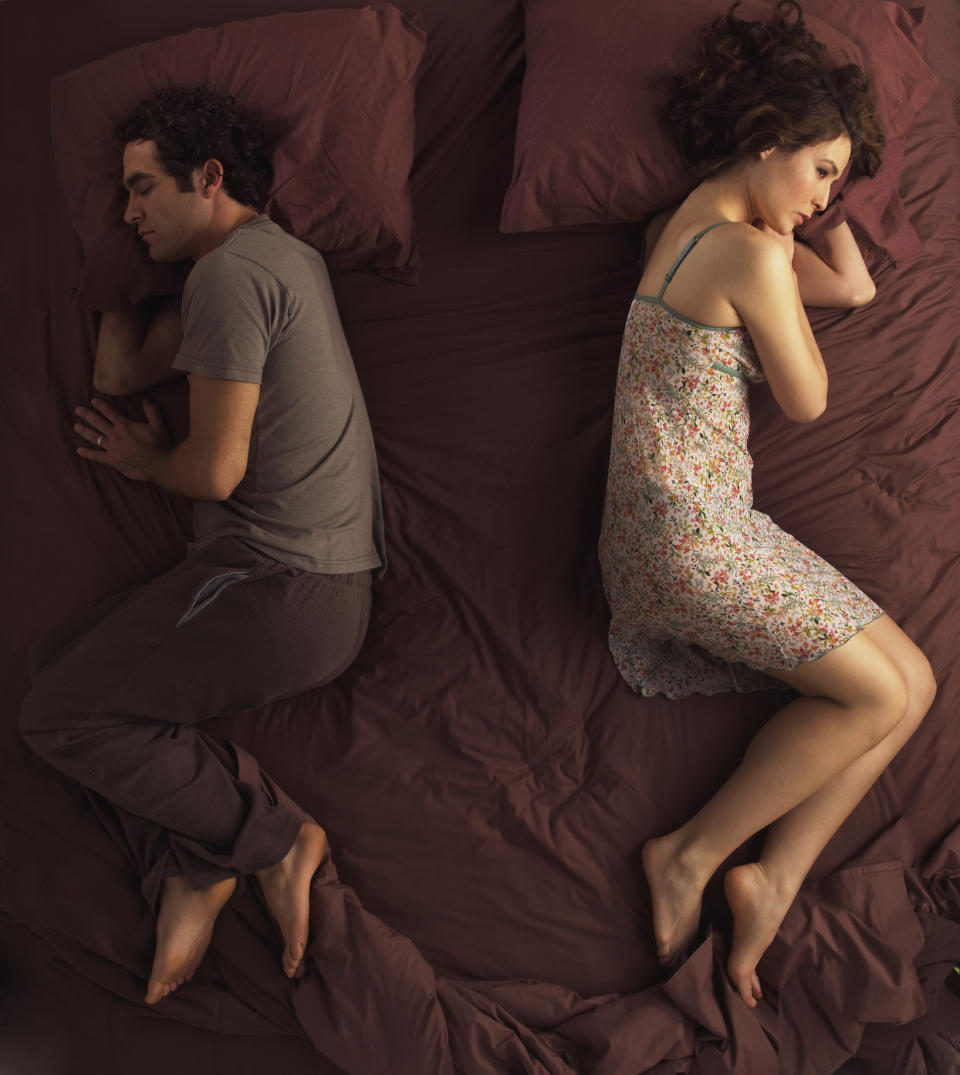 A man and a woman sleeping back-to-back on a bed, apparently in a disagreement or needing space