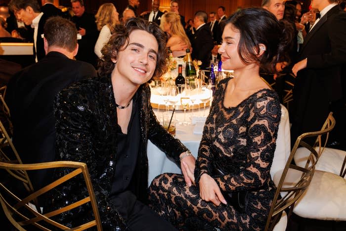 Timothée Chalamet and Selena Gomez sitting at a table, smiling at an event. Chalamet is wearing a sequin jacket, while Gomez is in a lacy black dress