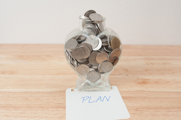 A jar of coins on top of a notecard that says "PLAN," on a wooden table.