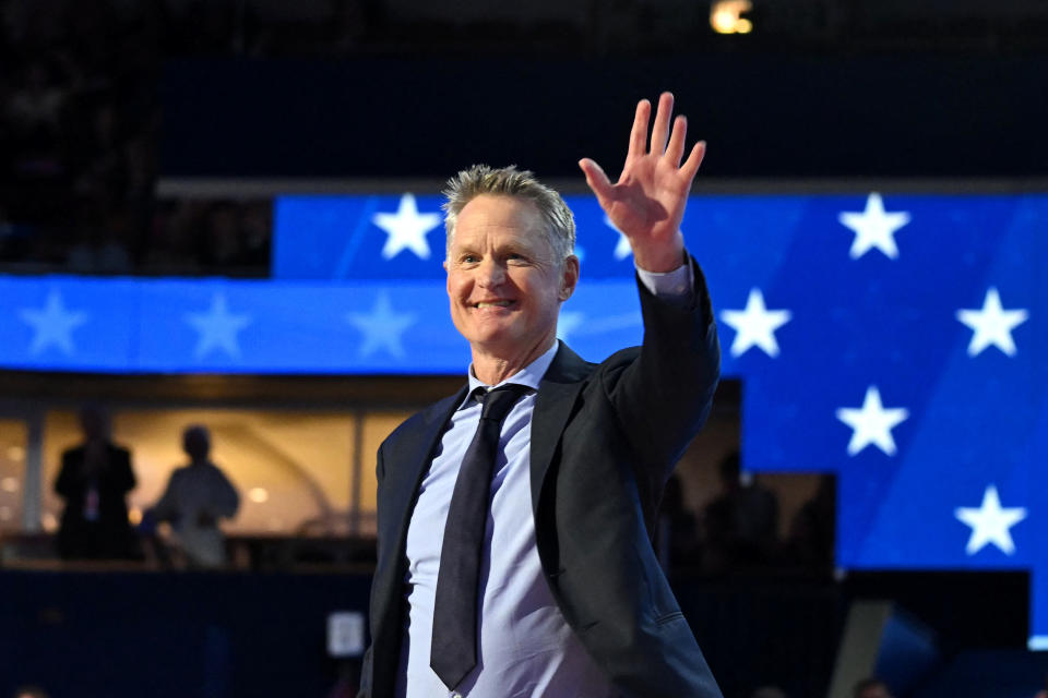 Steve Kerr winkt in Anzug und Krawatte vor einer Kulisse mit Sternen