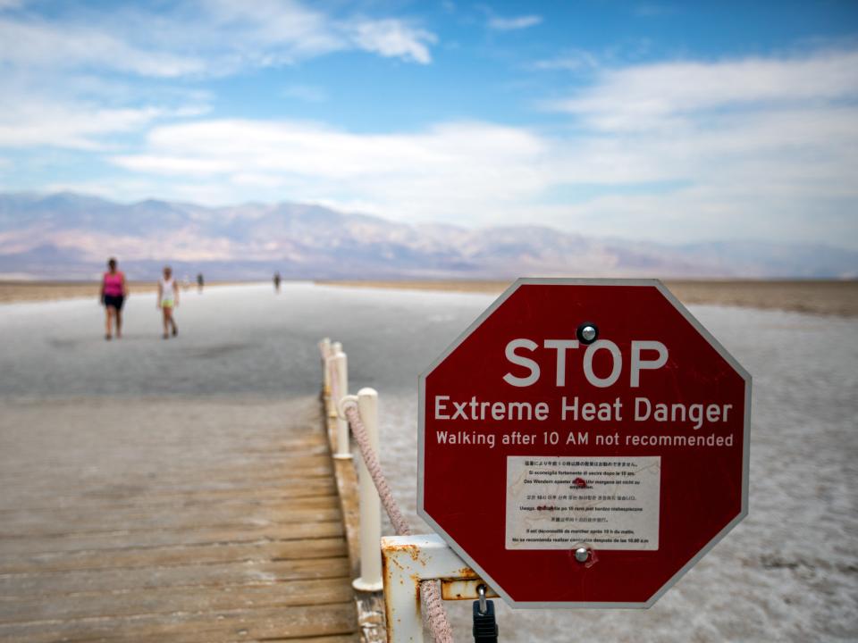 A red sign that reads "STOP, Extreme Heat Danger" next to a wooden walkway leading out into an expansive salt flat where tourists are walking.