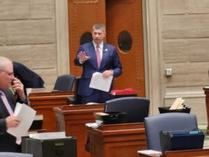 A man in a suit speaking in a legislative chamber. 