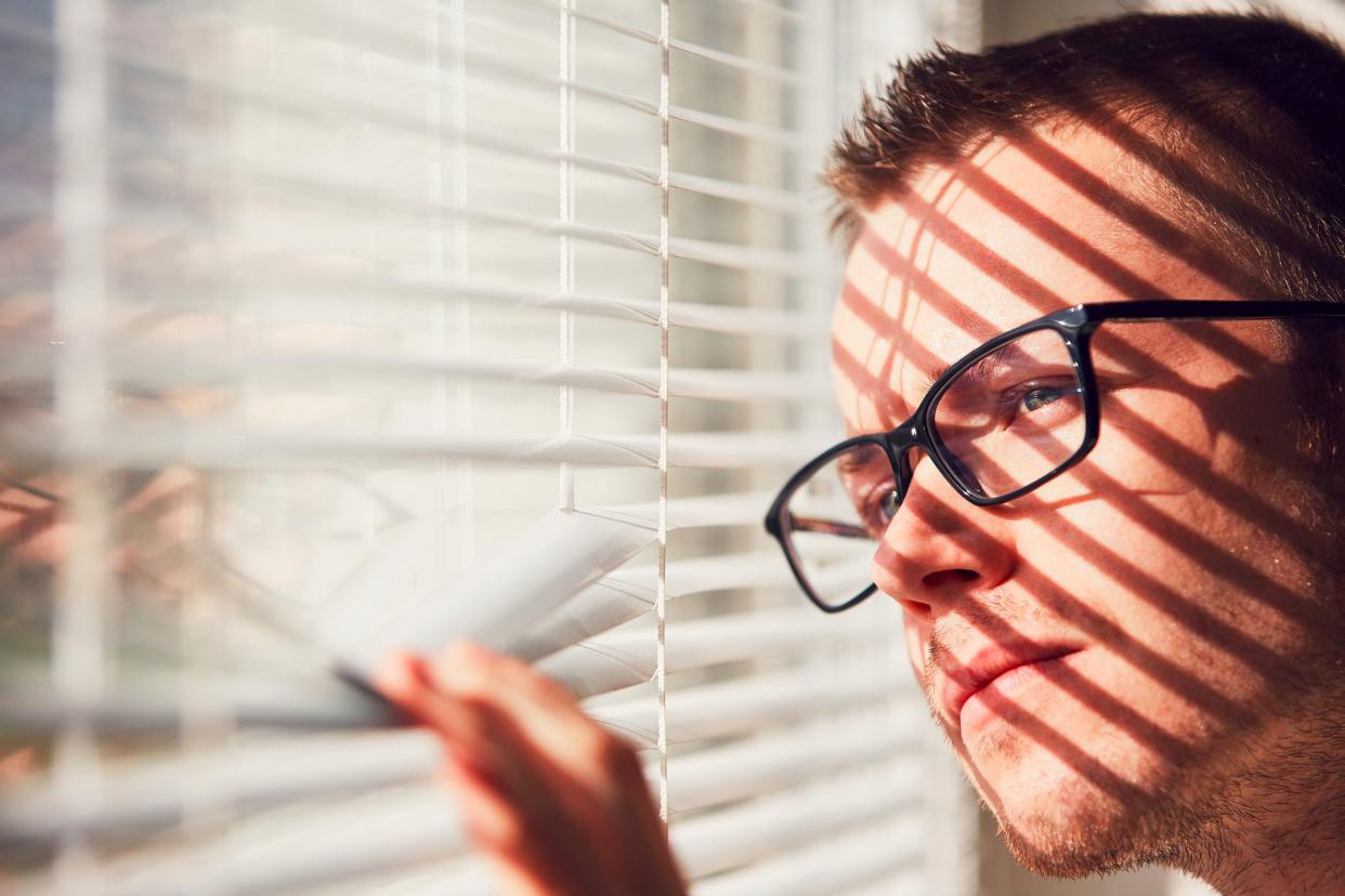 curious man, wearing glasses, bending a few blinds with his hand, peaking through the blinds out of his window