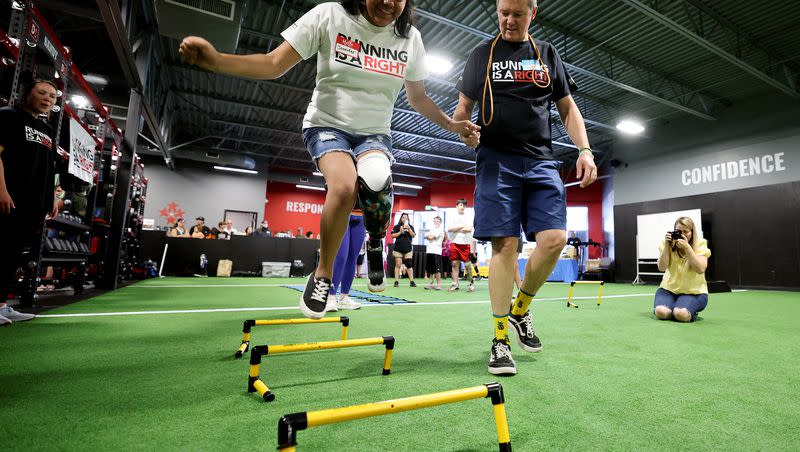 Jennifer Lozano is steadied by Mark Lange from Shriners as she jumps over some obstacles while joining other kids with prosthetic legs as they are assisted in reaching their running potential on Saturday, July 29, 2023. As part of their Running Is A Right program, the Dave McGillivray Finish Strong Foundation hosted a group of patients from Shriners Children’s Salt Lake City at a private event at D1 Training in Sandy, where specialists from Shriners Children’s Pediatric Orthotics and Prosthetics Services (POPS) helped the children with prosthetic legs reach their running potential.