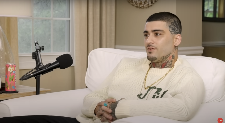 Zayn, wearing a sweater and gold chain, sits on a couch, speaking into a microphone during an interview