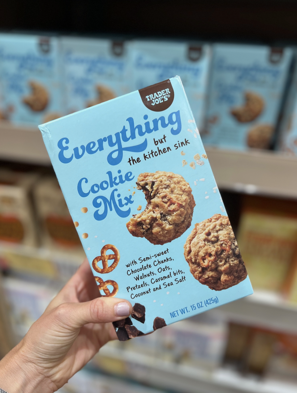 A hand holding a box of Trader Joe's Everything but the Kitchen Sink Cookie Mix. The box shows images of cookies, pretzels, and ingredients listed on it