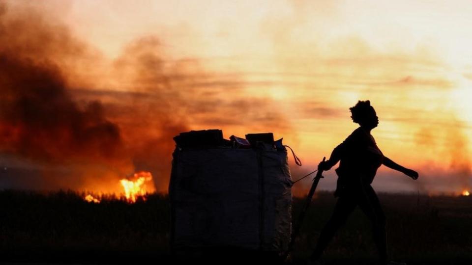 A woman pulls a cart with recyclables past field fires that take place as winter approaches, in Lenasia in southern Johannesburg, South Africa - April 22, 2024