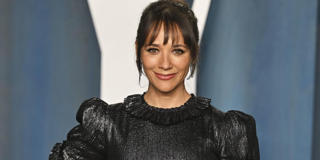 rashida jones, a woman stands with her right hand on her hip smiling at the camera, brown hair worn up with a fringe, wearing a black sparkly dress with puff sleeves