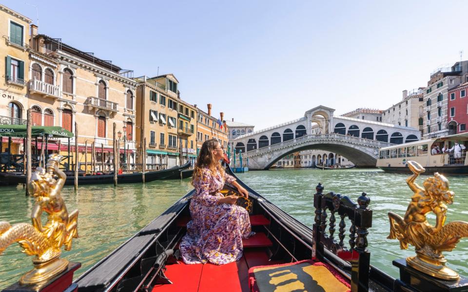 A young woman on a gondola enjoying a view of historic palazzi and the Rialto Bridge  - Getty