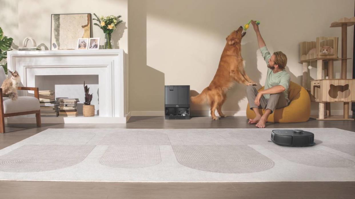  Lifestyle image of the eufy X10 Pro Omni cleaning carpet while a man plays with his dog next to the self-emptying station. 