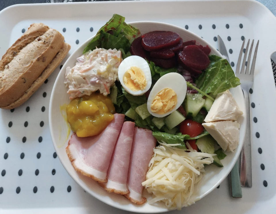 Plate of mixed salad with sliced ham, cheese, boiled egg, pickles, beetroot, coleslaw, and a bread roll on a tray with black dots