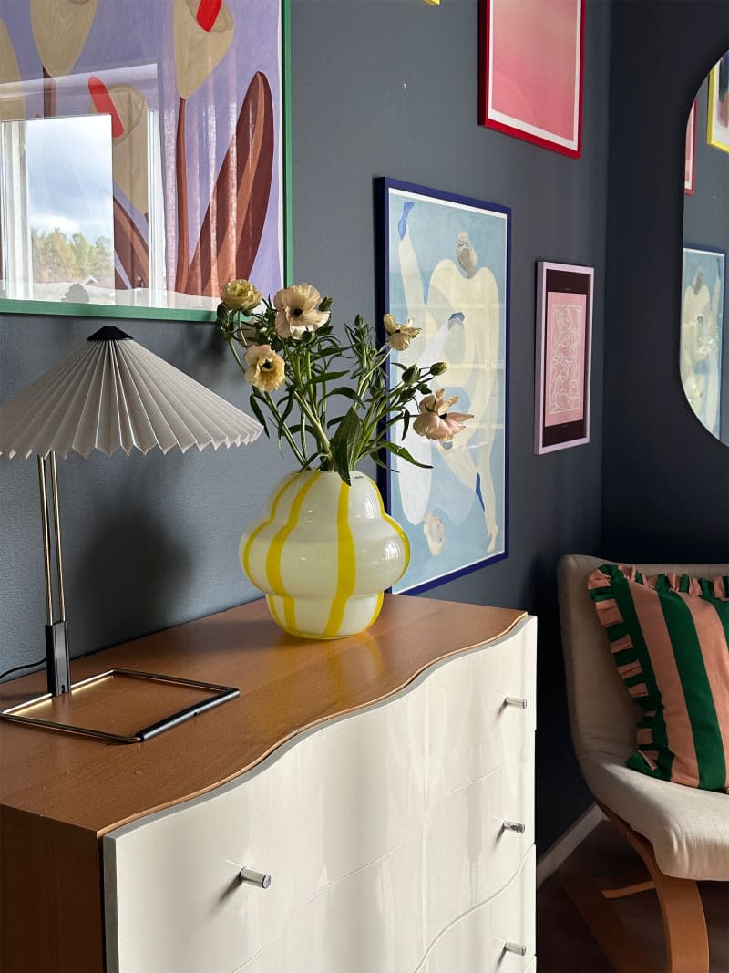 A multicolored vase sits on a dresser in a room filled with color and artwork.