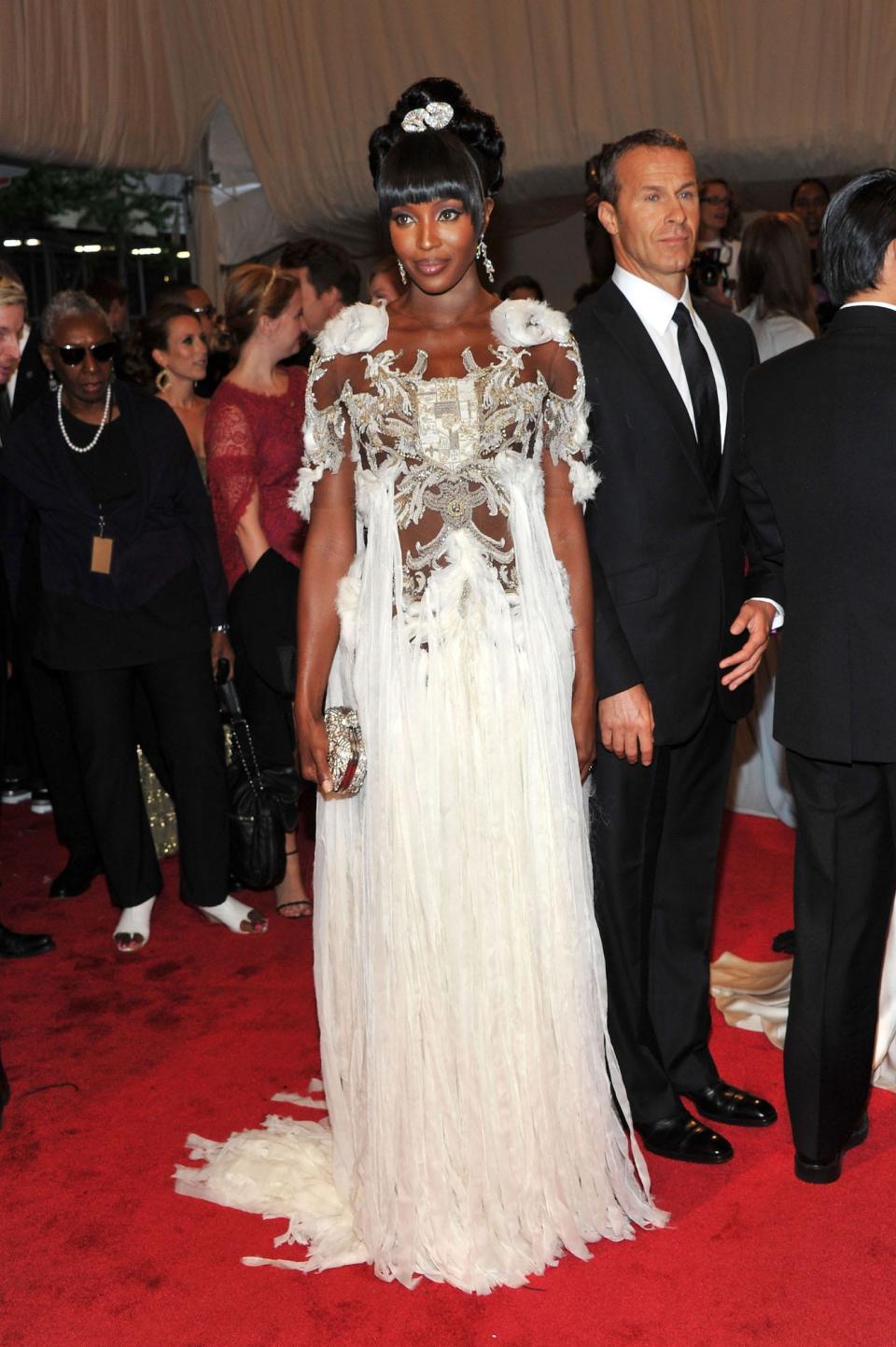 Naomi Campbell stands on the red carpet at the 2011 Met Gala wearing a flowing white dress decorated with a crest and silver detailing on the chest.