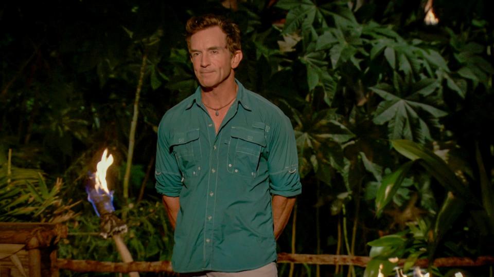 jeff probst stands with his hands behind his back in front of a torch on survivor