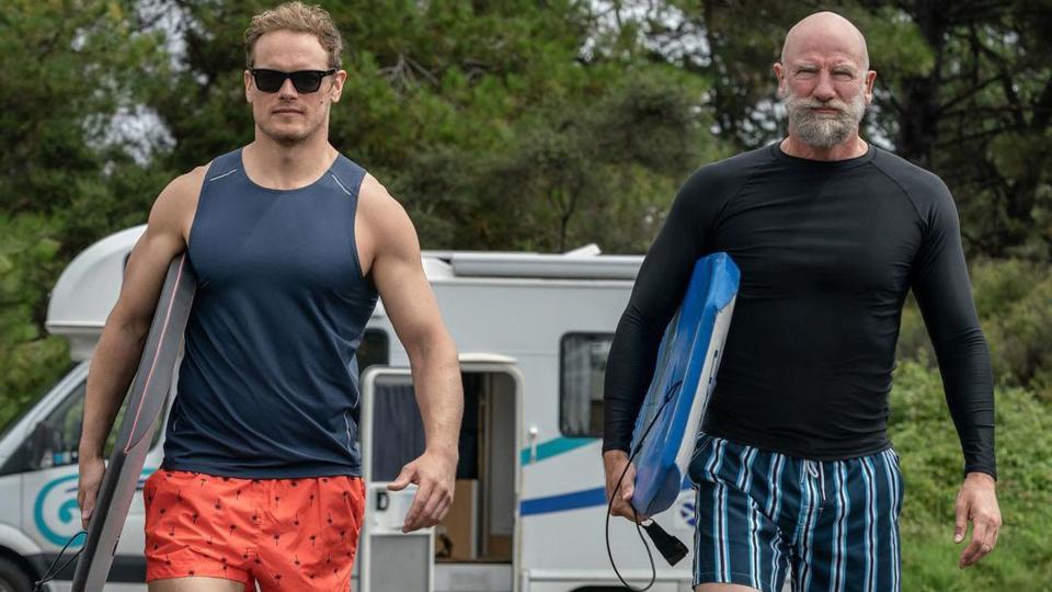 Sam Heughan and Graham McTavish carrying surf boards. 