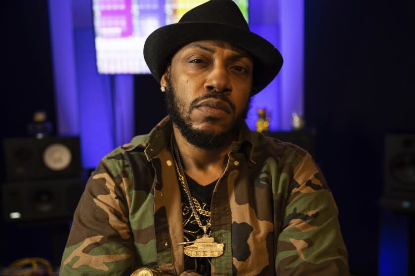 A beared man wearing a black hat, gold chains and camo jacket