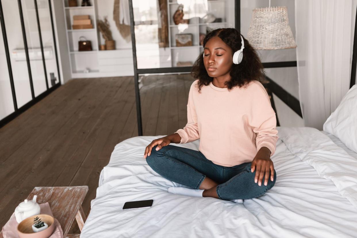 Relaxed African American Lady Meditating Listening To Music For Online Meditation Via Phone, Wearing Headphones And Closing Eyes Sitting In Lotus Position In Modern Bedroom At Home