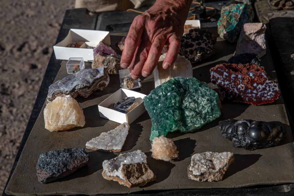 A multicolored assortment of rocks and minerals are arranged on a mat as a hand holds one of the specimens.